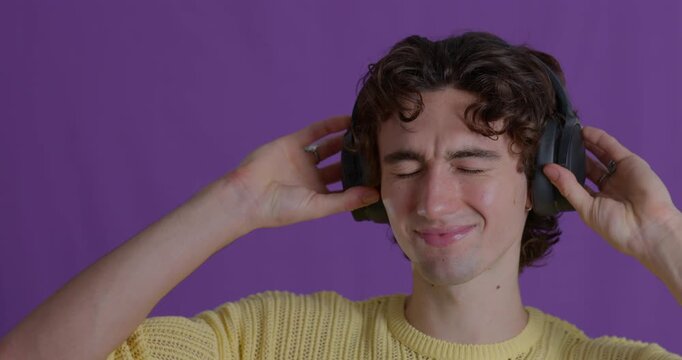 Close-up video clip of a smiling young adult male dancing while wearing wireless headphones, with his fingers pointing and eyes closed against a purple studio background. He has dark curly hair
