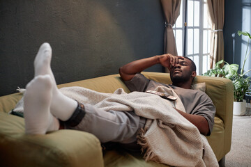 Black man in his twenties lying on sofa with electronic ankle monitor visible, resting under...