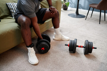 Black young adult man sitting on sofa wearing electronic ankle monitor holding dumbbell with...