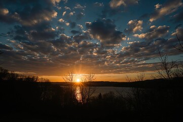 Breathtaking evening scene of a calm lake framed by leafless trees, bathed in a golden light with vibrant clouds overhead.