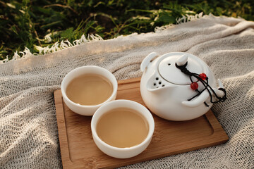 Teapot with two cups filled with tea on a wooden tray, placed outdoors on a rustic fabric. Close view