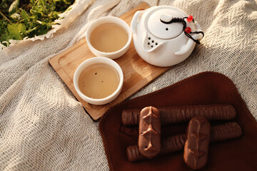 Teapot with cups of tea on a tray, next to chocolate biscuits on a brown cloth. Picnic for two people outdoor.
