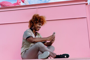 Smiling young African American man with curly ginger hair sitting cross-legged outdoors, holding smartphone with both hands, enjoying digital connection and modern lifestyle against pink wall.