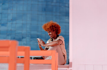 Smiling young African American man with curly ginger hair sitting outdoors, holding smartphone in both hands, enjoying connection, instant messaging, and positive digital interaction.