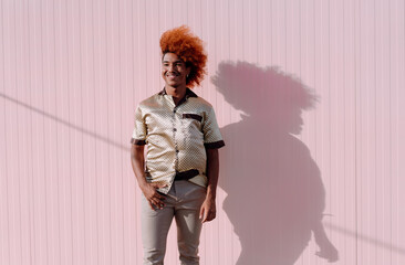 Young African American man with curly ginger hair standing outdoors against pink wall, smiling with...