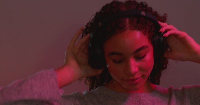 Video clip of a young adult female smiling and dancing in the studio while listening to music through wireless headphones. She has brunette curly hair and she is illuminated by colourful neon disco li