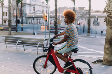 Side view of young man with afro hairstyle riding rental bicycle. Stock photo concept of...