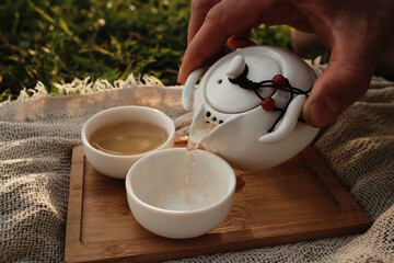 Hand pouring hot tea from a white teapot into a small porcelain cup outdoors on a wooden tray. Picnic in Chinese style for two people outdoor