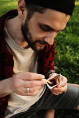 Young bearded man in plaid shirt dipping French fries into sauce cup during picnic in a park