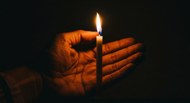 Holding a single lit candle in hand with warm glow against dark background, symbolizing hope, remembrance, or peace. - Powered by Adobe