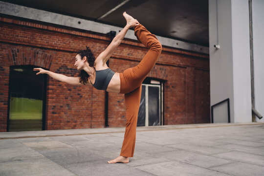 Athletic woman practicing yoga dancer pose on city pavement, extending arm forward with grace. Conceptual photo symbolizing empowerment, focus, resilience, balance, and mindfulness in modern life.
