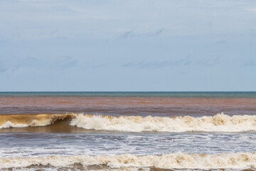Waves crashing on a sandy beach with a cloudy sky and a mix of brown and blue water in the ocean