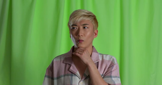 Video clip of a young adult male pondering in the studio, looking around with one hand to his chin, before smiling at the camera. He has bleached hair and is wearing casual clothing.