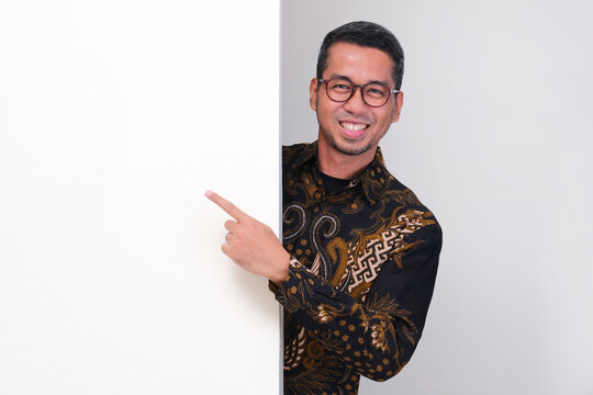 Indonesia school teacher peeking behind a wall and pointing on it with happy face