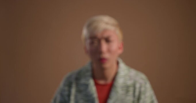 Video clip of a young adult male looking for and putting on his glasses in the studio. The clip begins blurred and comes into focus as he puts on his glasses. He has bleached hair and is wearing casua
