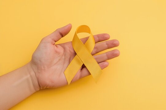 Support for individuals with illness shown by hand holding yellow ribbon against yellow backdrop, symbolizing Suicide Prevention Day, Sarcoma Cancer, and Childhood Cancer Awareness Month