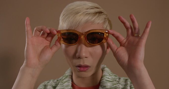 Close-up video clip of a young adult male checking his appearance close to the camera in the studio. He has bleached hair and is wearing sunglasses and casual clothing.