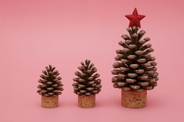 Three pine cones of varying sizes placed on cork stands on a pink background, mimicking miniature trees. A big pine cone decorated with a red star topper, highlighting a holiday-themed DIY project