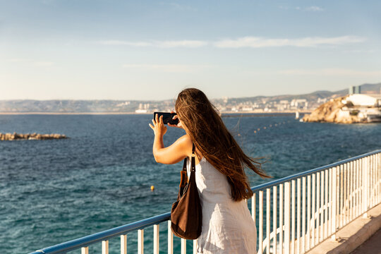 Jeune femme prenant une photo avec son smartphone, vue de trois quart arri&egrave;re, les cheveux longs au vent, se tenant debout sur la corniche Kennedy surplombant la mer, Marseille, Sud de la France