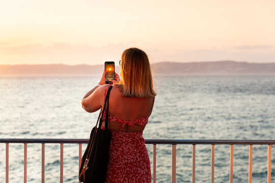 Femme prenant une photo du coucher du soleil sur la mer avec son smartphone, vue de trois quart arri&egrave;re, portant une robe d'&eacute;t&eacute;, se tenant debout sur la corniche Kennedy, Marseille, Sud de la France
