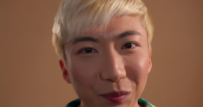 Close-up video clip of a young adult male messing around in the studio, gesturing playfully towards the camera and laughing. He has bleached hair and is wearing a multicoloured shirt.