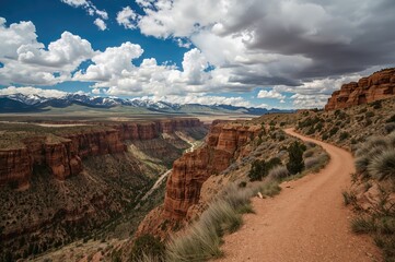 Fototapeta premium A serpentine trail beside towering sandstone walls with expansive cloudy skies above and distant snow-capped mountains enhancing the stunning vista.