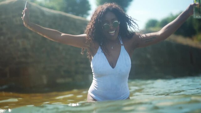 A black woman is holding a cell phone and a drink while standing in a pool. She is smiling and she is having fun
