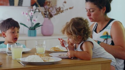 Mother having lunch with two children at home, toddler seated at table holding fork while mother assists, casual family mealtime moment with rice, chicken, and juice on wooden table - Powered by Adobe