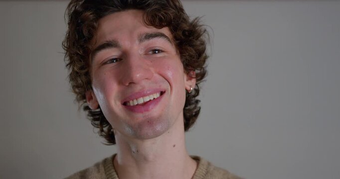 Close-up video clip of a young adult male smiling at the camera in the studio. He has dark curly hair and is wearing a casual sweatshirt.