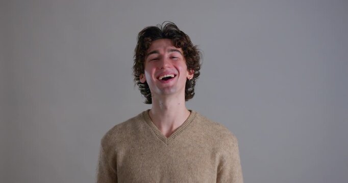 Medium video clip of a young adult male laughing towards the camera in the studio. He has dark curly hair and is wearing a casual sweatshirt.