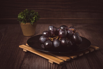 Bunch of fresh purple grapes on a black plate, placed on a wooden base, with a rustic background. Natural style image, ideal for concepts of healthy eating, fruits, nutrition and gastronomy.