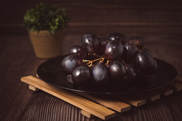 Bunch of fresh purple grapes on a black plate, placed on a wooden base, with a rustic background. Natural style image, ideal for concepts of healthy eating, fruits, nutrition and gastronomy.