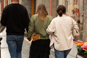 Three friends walking together down a narrow city street, one wearing a green jacket and holding hands with another.