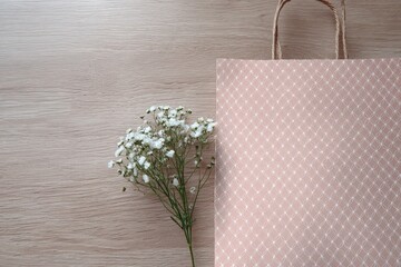 A light pink paper gift bag sits on a light beige wooden surface beside a small bouquet of white flowers.