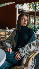 Elegant young woman wearing a beige trench coat and black turtleneck, seated at a round table in a cozy outdoor cafй with rattan chairs and white curtains