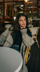 Obraz premium Stylish young woman in a trench coat and black turtleneck sitting alone at a table in a cozy outdoor cafe, looking thoughtful and composed