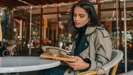 Obraz premium Elegant young woman in a beige trench coat and black turtleneck sitting at a round table in a cafй, reading an old book with deep concentration