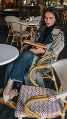 Elegant young woman in a beige trench coat and black turtleneck sitting at a round table in a caf&eacute;, reading an old book with deep concentration