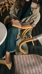 Elegant young woman in a beige trench coat and black turtleneck sitting at a round table in a cafй, reading an old book with deep concentration