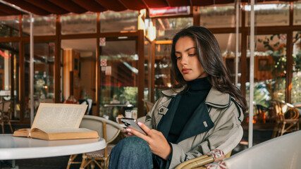 Fototapeta premium Young woman in trench coat sitting at outdoor cafe table with vintage book and smartphone. Autumn mood with elegant style and cozy ambience