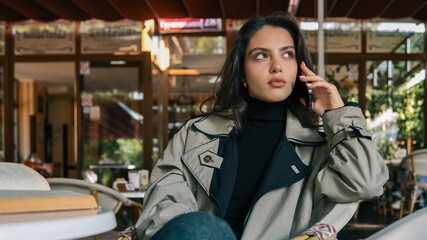 Elegant young woman in a trench coat sitting at an outdoor cafe, holding smartphone to her ear, engaged in conversation. Autumn scene with a cozy, relaxed mood