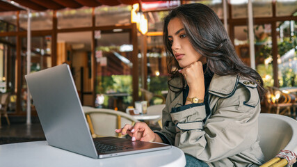 Young woman in trench coat sitting at outdoor cafй, resting her chin on hand while looking at...