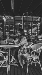 Side view of a young woman in a beige trench coat sitting at a white round table with a laptop in an outdoor cafй. She is focused and thoughtful, surrounded by empty chairs and soft natural light