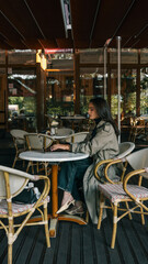 Side view of a young woman in a beige trench coat sitting at a white round table with a laptop in an outdoor cafй. She is focused and thoughtful, surrounded by empty chairs and soft natural light