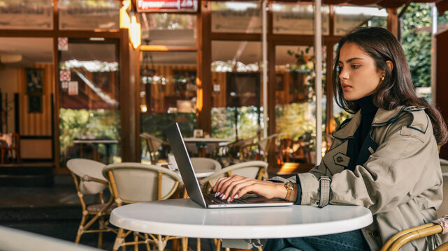 Confident young woman in a beige trench coat resting her head on hand while using a laptop at a white outdoor cafй table. Looking at camera with calm, thoughtful expression