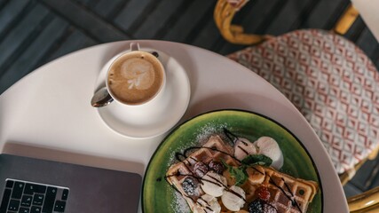 Top view of delicious waffles with berries, banana, ice cream scoops, chocolate syrup, and a cappuccino with latte art served on a white table next to a laptop in a cozy café setting
