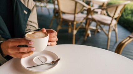 Close-up of woman’s hands in trench coat gently holding a cup of cappuccino with latte art,...