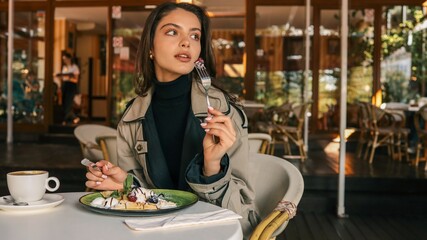 Young stylish woman holding a fork with raspberry, enjoying a sweet waffle dessert with coffee at an elegant outdoor café