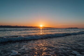 Stunning sunset over serene ocean waves with mountains in the background, vibrant orange hues lighting up the blue sky, captured with long exposure.