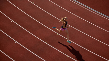 Aerial view of female athlete running on red track during sunny day. Concept of overhead perspective, athlete sprint motion, rhythm of speed, contrast of body and shadow, dynamic track environment.